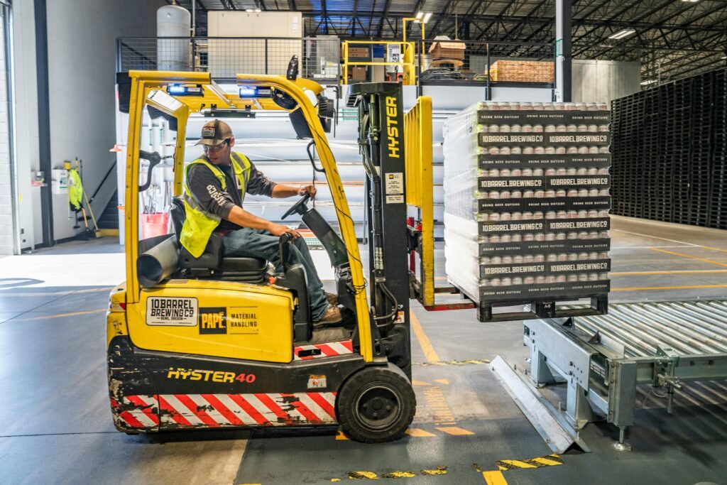 A warehouse worker maneuvers a forklift to transport crates for brewing company storage.