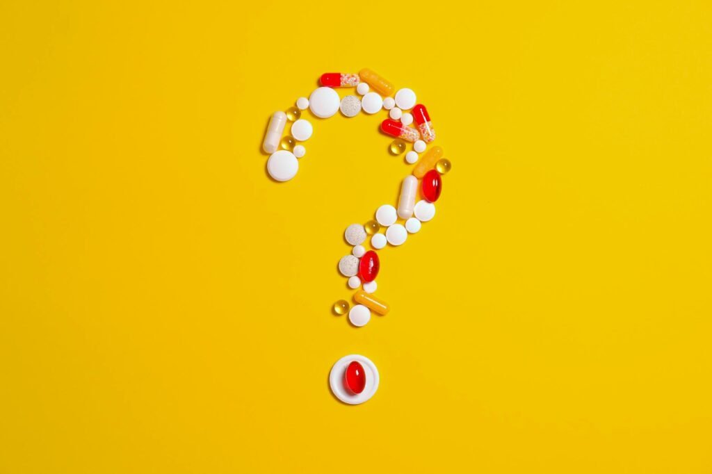 Assorted pills and capsules arranged in a question mark on a bright yellow background symbolizing medication inquiries.