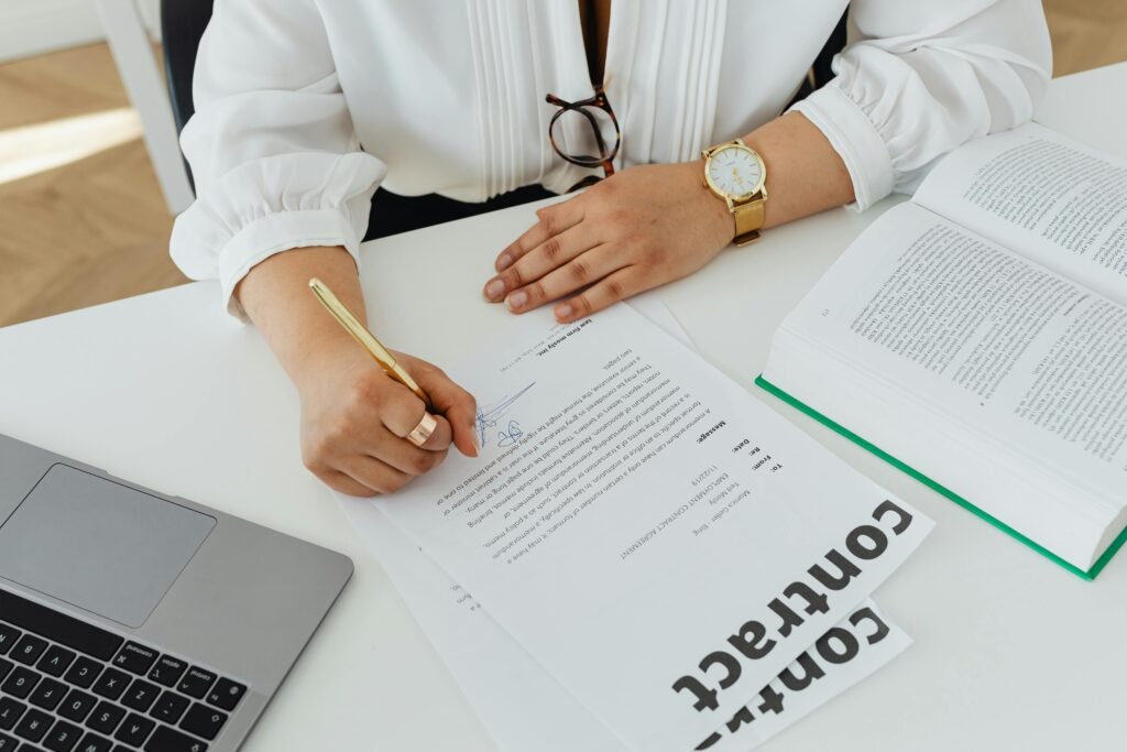 Close-up of a businesswoman signing a contract at her desk, with a laptop and book.